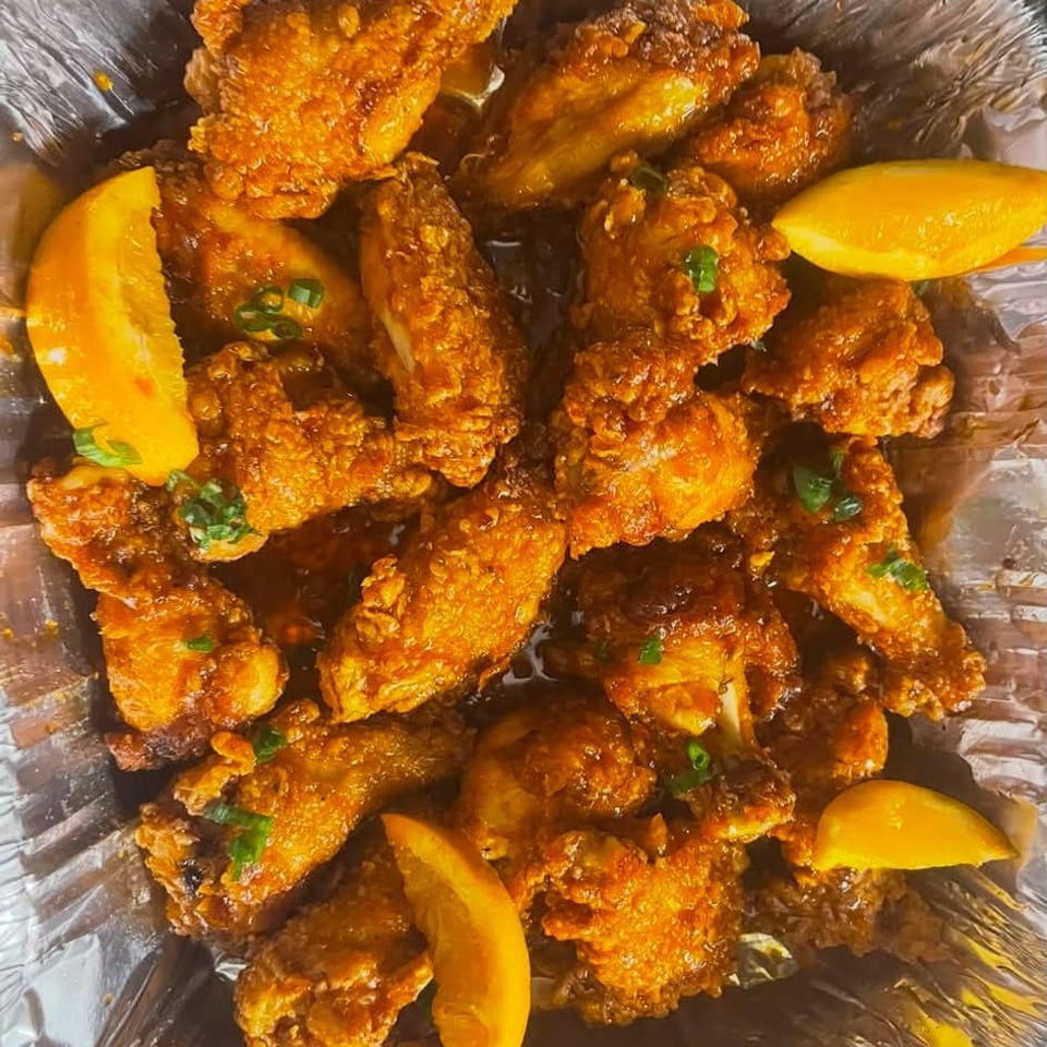 Crispy sauced chicken wings garnished with green onions and orange wedges in a foil serving tray.