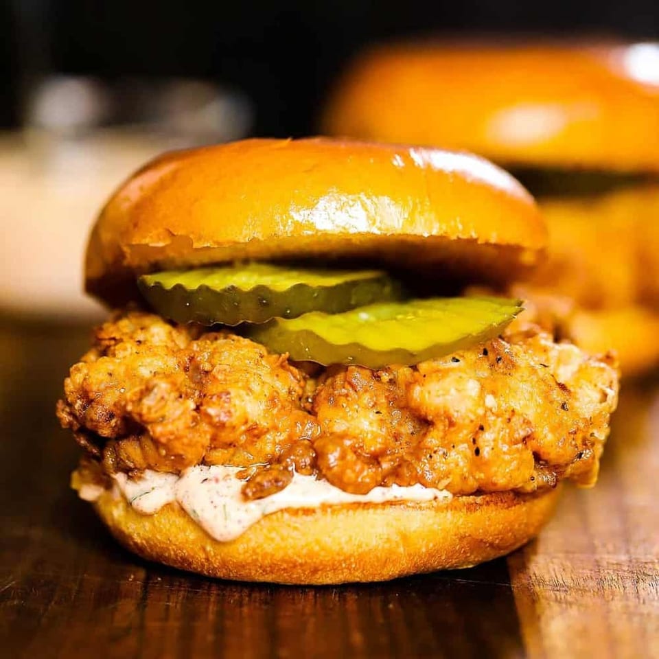 Crispy fried chicken sandwich on a glossy bun with pickles and creamy sauce, served on a wooden table.