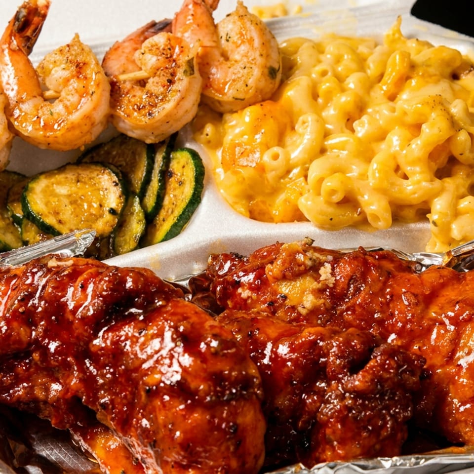 Takeout container with barbecue wings, grilled shrimp skewers, macaroni and cheese, and zucchini.